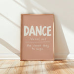 A framed typographic wall art print leaning against a white wall on a light wood floor. The print has a dusty blush pink background with white text reading “DANCE /dɑːns/, verb” and “the closest thing to magic.
