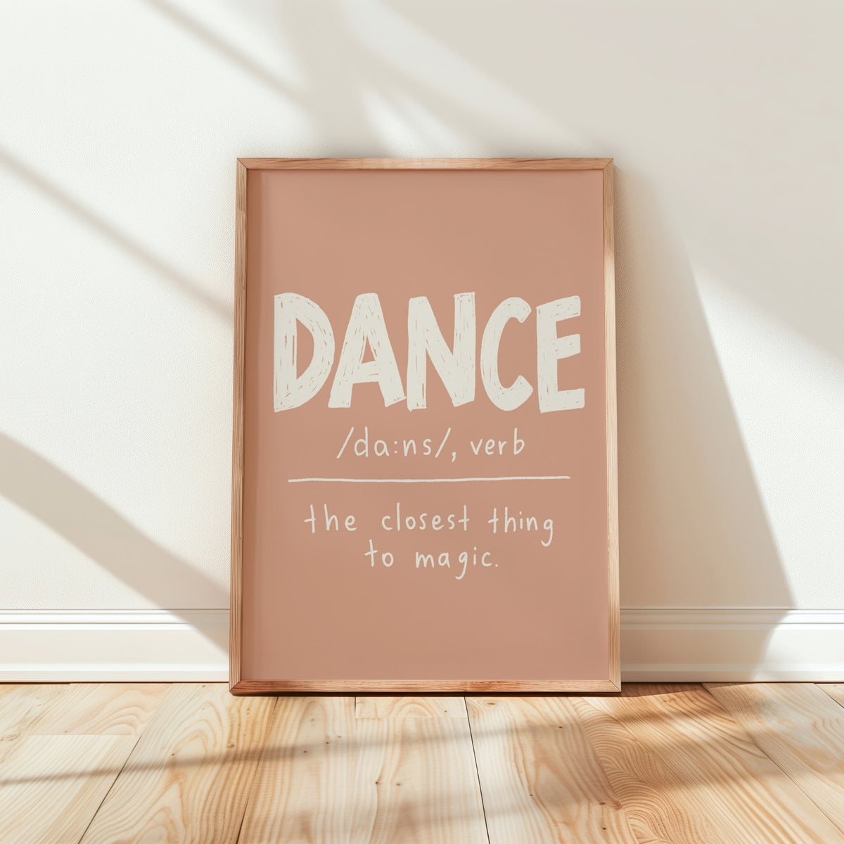 A framed typographic wall art print leaning against a white wall on a light wood floor. The print has a dusty blush pink background with white text reading “DANCE /dɑːns/, verb” and “the closest thing to magic.