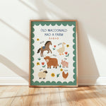 A framed children's educational poster titled 'OLD MACDONALD HAD A FARM' leaning against a white wall on a light wood floor. The poster features a green border, a cream center, and illustrations of farm animals including a horse, goose, pig, sheep, chicken, and cow, each labeled with its sound.