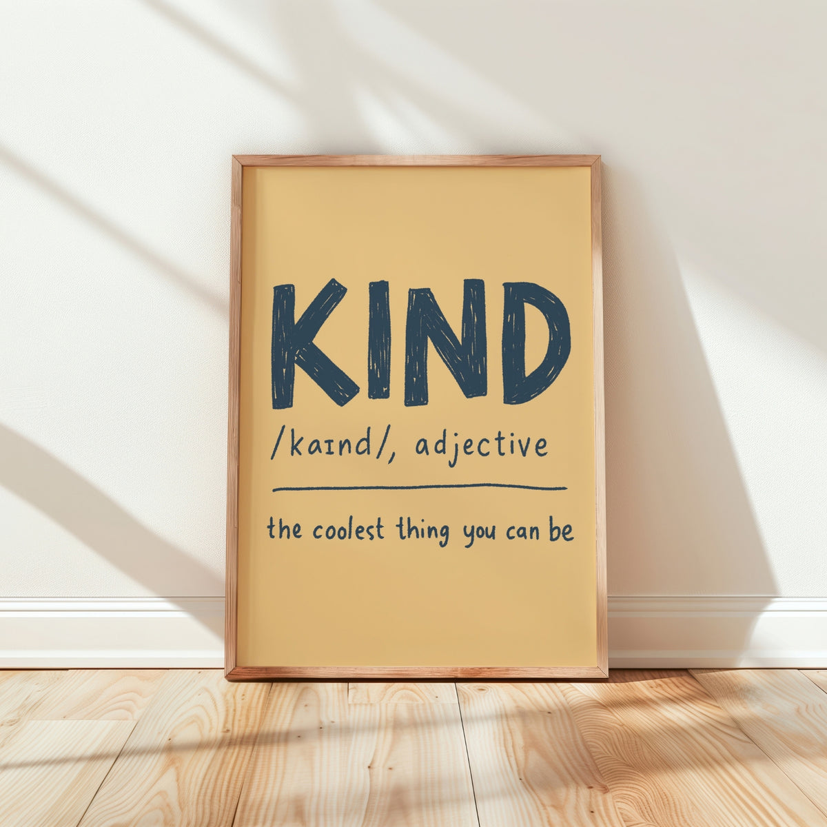 A framed typographic wall art print leaning against a white wall on a light wood floor. The print has a warm cream background with dark navy text reading “KIND /kaɪnd/, adjective” and “the coolest thing you can be.