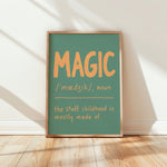 A framed typographic wall art print leaning against a white wall on a light wood floor. The print has a muted teal-green background with large orange text reading “MAGIC” and smaller orange text below defining the word and the phrase “the stuff childhood is mostly made of.