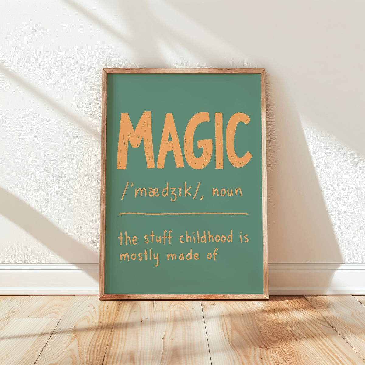A framed typographic wall art print leaning against a white wall on a light wood floor. The print has a muted teal-green background with large orange text reading “MAGIC” and smaller orange text below defining the word and the phrase “the stuff childhood is mostly made of.