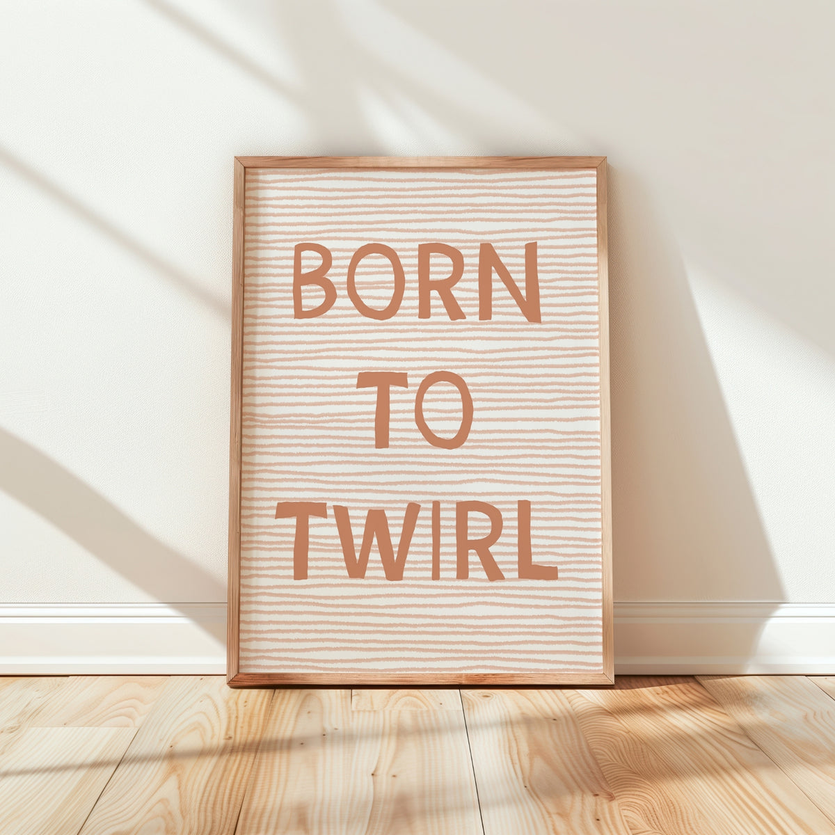 A framed typographic wall art print leaning against a white wall on a light wood floor. The print has a natural wood frame, a cream background with fine horizontal wavy lines, and the words 'BORN TO TWIRL' in large peach-pink letters.