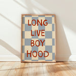 A framed poster with a beige and blue-gray checkerboard pattern and the words 'LONG LIVE BOY HOOD' in bold red lettering, leaning against a white wall on a light wood floor.