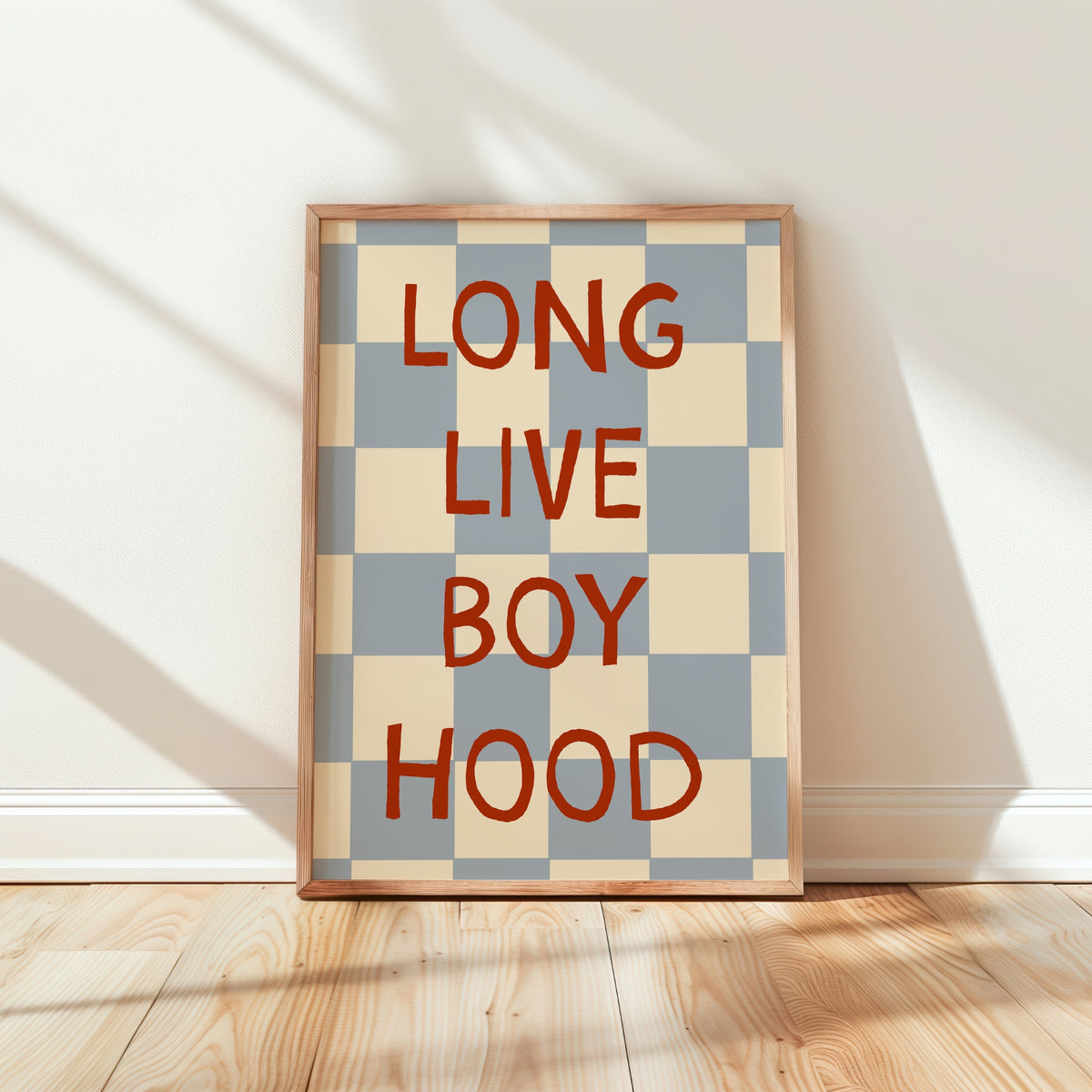 A framed poster with a beige and blue-gray checkerboard pattern and the words 'LONG LIVE BOY HOOD' in bold red lettering, leaning against a white wall on a light wood floor.