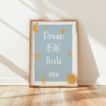 A framed children's wall art print leaning against a white wall on a light wood floor. The print has a pale blue background with the phrase 'Dream BIG little one' in white script, decorated with gold stars, a crescent moon, and a sun, in a light wood frame.