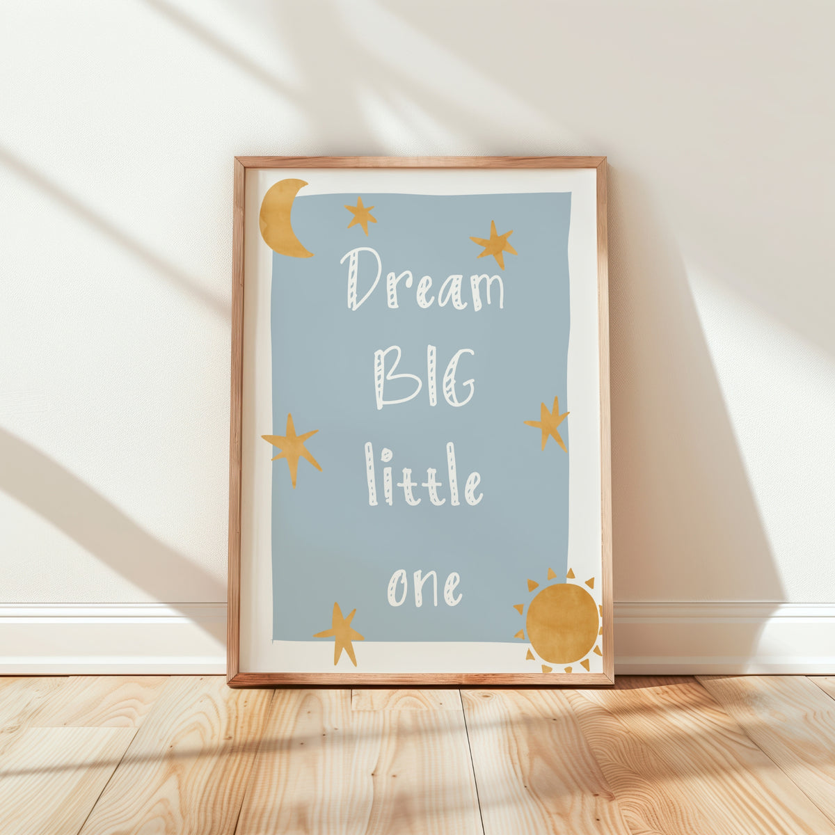 A framed children's wall art print leaning against a white wall on a light wood floor. The print has a pale blue background with the phrase 'Dream BIG little one' in white script, decorated with gold stars, a crescent moon, and a sun, in a light wood frame.