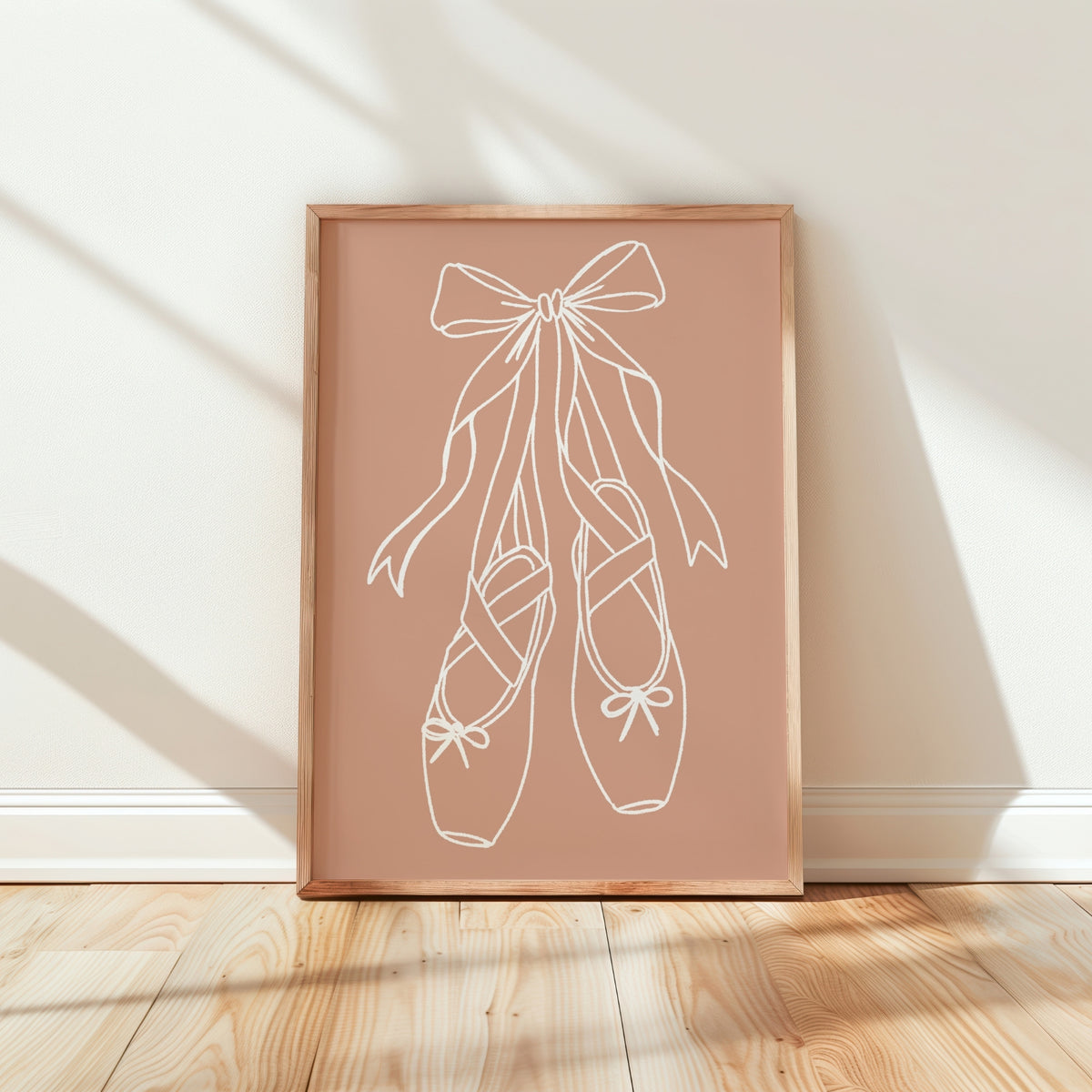 A framed minimalist art print featuring a white line drawing of a ballerina's tutu with a small heart on the chest, set against a dusty rose background. The print is housed in a light natural wood frame and is leaning against a white wall on a light wood floor, with soft diagonal shadows cast across the scene.