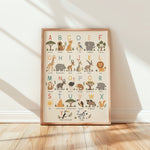 A framed children's alphabet art print featuring illustrated safari animals and letters, leaning against a white wall on a light wood floor with soft window shadows.