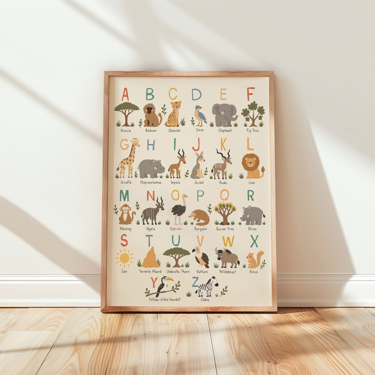 A framed children's alphabet art print featuring illustrated safari animals and letters, leaning against a white wall on a light wood floor with soft window shadows.