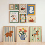 A set of nine framed country-themed art prints arranged on a white wall above a light wood floor, including illustrations of a cow, flowers in a watering can, a horse, a tractor, a sheep, and farm animals.