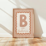 A framed vertical nursery print leaning against a white wall on a light wood floor. The print features a large letter B inside a stamp-like border with the words 'is for Ballerina' in a warm peach and cream color palette.