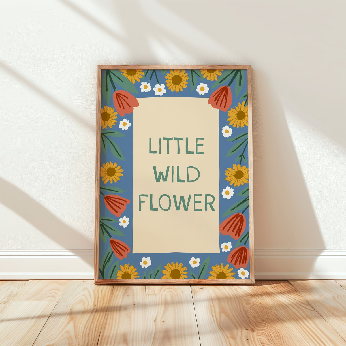 A framed art print with the text 'LITTLE WILD FLOWER' in green on a cream background, surrounded by a blue border with yellow sunflowers, white daisies, and orange-red tulips. The frame is leaning against a white wall on a light wood floor with soft diagonal shadows.