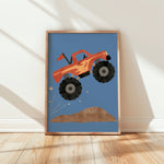 A framed art print featuring a stylized orange monster truck jumping over a brown hill on a blue background, leaning against a white wall on a light wood floor.