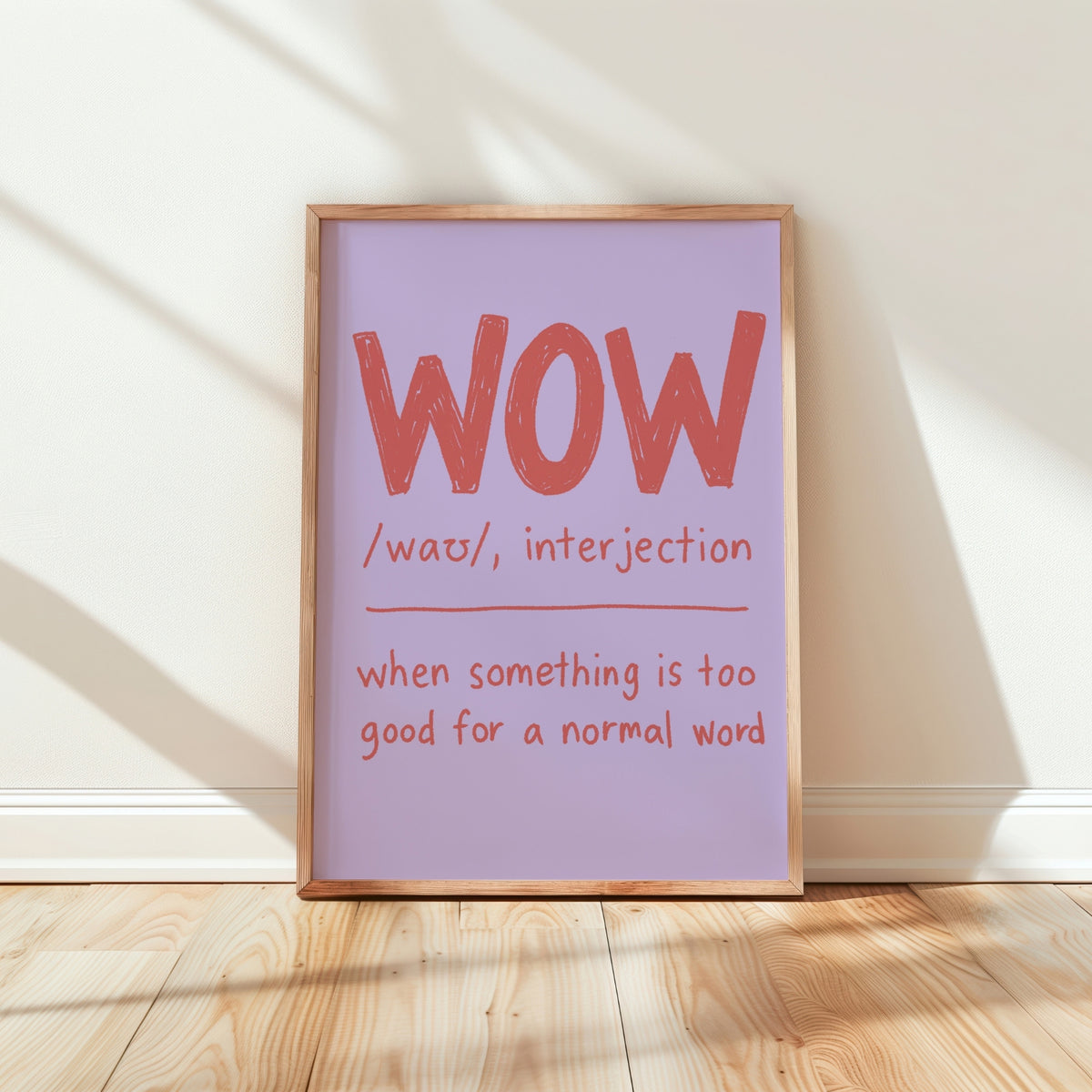 A framed typographic wall art print leaning against a white wall on a light wood floor. The print has a lavender background with large coral-red text reading “WOW” and smaller text below defining the word, plus a light natural wood frame.