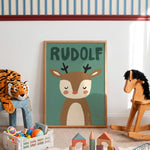A framed personalized children's art print featuring a cartoon deer with the name 'RUDOLF' above it, displayed in a styled nursery room with toys and a rocking horse.