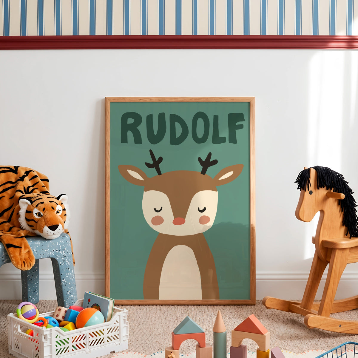A framed personalized children's art print featuring a cartoon deer with the name 'RUDOLF' above it, displayed in a styled nursery room with toys and a rocking horse.