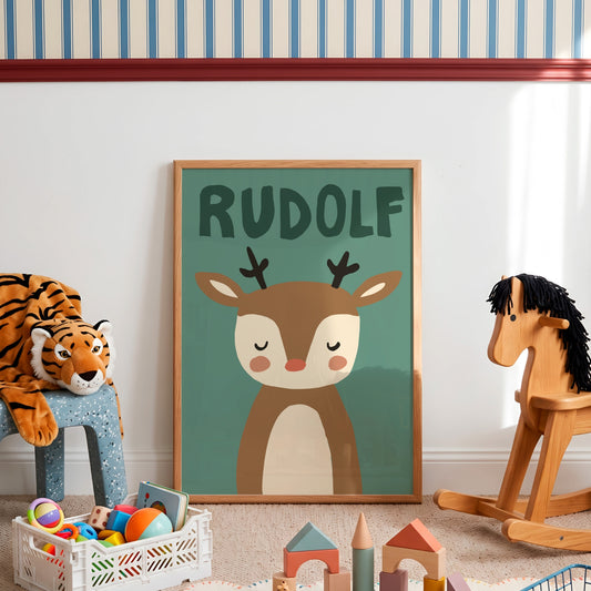 A framed personalized children's art print featuring a cartoon deer with the name 'RUDOLF' above it, displayed in a styled nursery room with toys and a rocking horse.