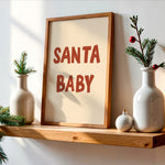 A framed wall art print with the words “SANTA BABY” in large reddish-brown letters on a cream background, leaning on a wooden shelf beside white ceramic vases with pine branches and small ornaments.