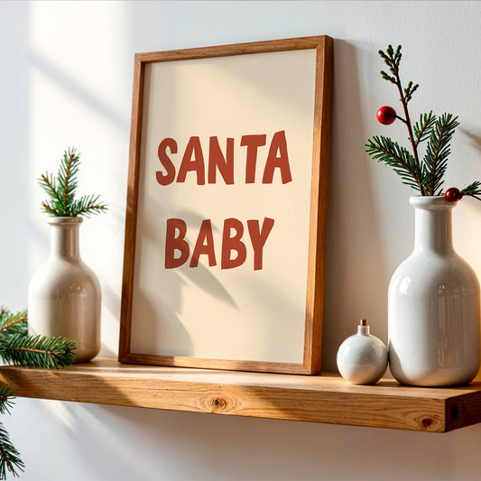 A framed wall art print with the words “SANTA BABY” in large reddish-brown letters on a cream background, leaning on a wooden shelf beside white ceramic vases with pine branches and small ornaments.