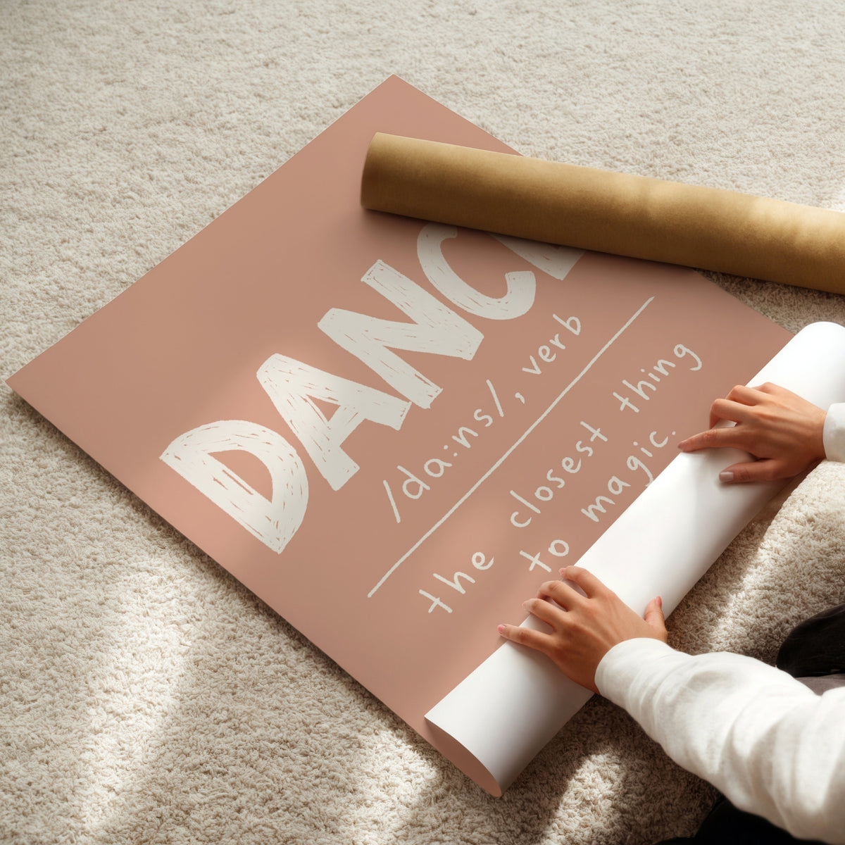 A large rolled poster or print in dusty pink with white text reading 'DANCE /dæns, verb the closest thing to magic' is being unrolled on a beige carpet. A person's hands and arms are visible holding the roll, and a brown cardboard mailing tube lies nearby.