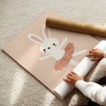 A person is unrolling a large pastel pink wall mural featuring a white bunny ballerina with a dusty pink tutu, placed on a beige carpet. A brown cardboard shipping tube lies nearby.