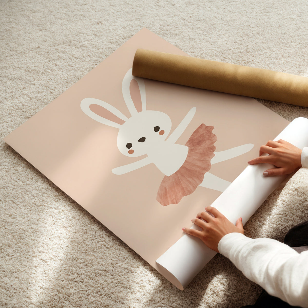 A person is unrolling a large pastel pink wall mural featuring a white bunny ballerina with a dusty pink tutu, placed on a beige carpet. A brown cardboard shipping tube lies nearby.