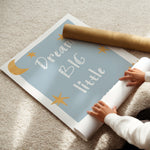 A person is unrolling a large decorative wall poster or mural on a beige carpet. The poster has a light blue background with white text reading 'Dream Big little' and gold star and moon illustrations.