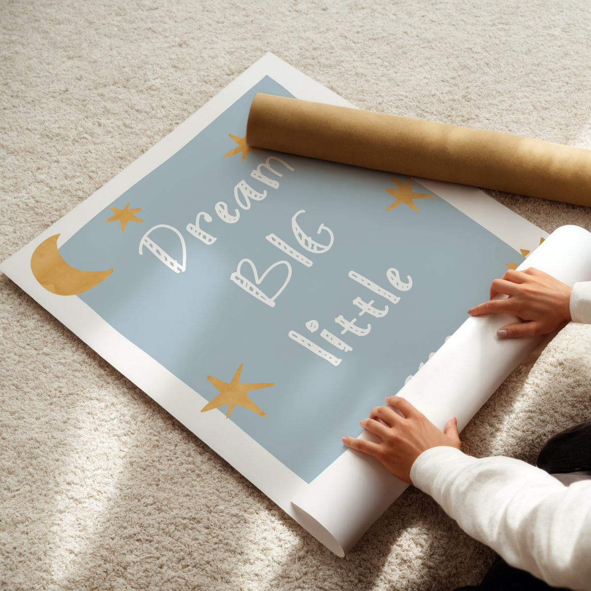 A person is unrolling a large decorative wall poster or mural on a beige carpet. The poster has a light blue background with white text reading 'Dream Big little' and gold star and moon illustrations.