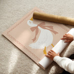 A person unrolls a large decorative wall mural or poster featuring a white duck with a tan bow and yellow beak/legs on a soft blush background. The mural is partially rolled, with a brown cardboard tube placed on top, set on a beige carpet.