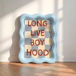 A square decorative wall art piece with a wavy light blue frame, leaning against a white wall on a wood floor. The center shows a gray and cream checkerboard pattern with the words 'LONG LIVE BOY HOOD' in red block letters.