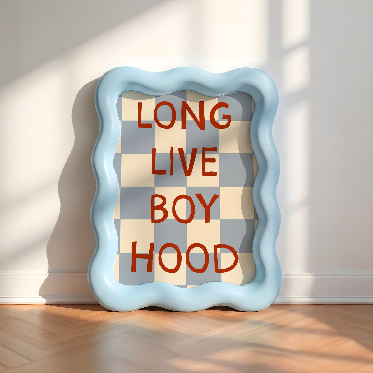 A square decorative wall art piece with a wavy light blue frame, leaning against a white wall on a wood floor. The center shows a gray and cream checkerboard pattern with the words 'LONG LIVE BOY HOOD' in red block letters.