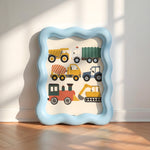 A wavy light blue picture frame with a children's construction vehicle print, leaning against a white wall on a wooden floor with soft window shadows.