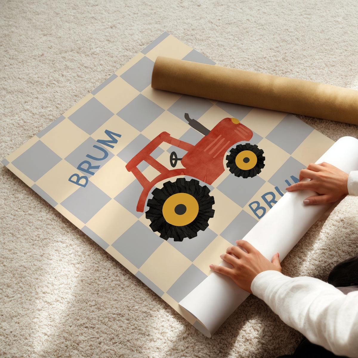 A person is unrolling a personalized children's wall decal featuring a red tractor on a beige and gray checkered background with the name 'BRUM'. A brown cardboard shipping tube lies nearby on a beige carpet.