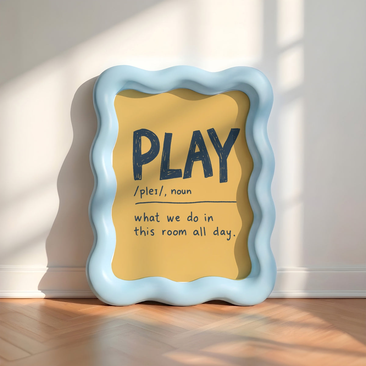 A wavy light blue picture frame leaning against a white wall on a wooden floor, with a yellow insert that reads “PLAY /pleɪ/, noun what we do in this room all day.” Sunlight casts shadows across the wall and floor.