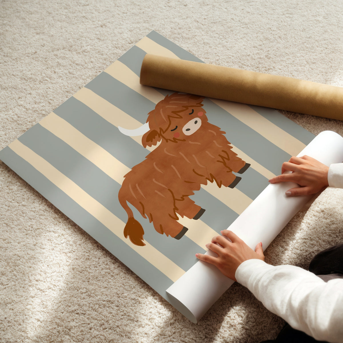 A person is unrolling a sheet of wallpaper on a beige carpet. The wallpaper features a design of alternating beige and muted blue-gray vertical stripes with a central illustration of a fluffy brown Highland cow.