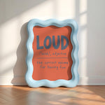 A decorative square sign with a wavy light blue frame leans against a white wall on a wood floor. The sign has a coral-orange center with the word “LOUD” in large gray letters and smaller handwritten-style text underneath reading “/lauid, adjective” and “the correct volume for having fun.