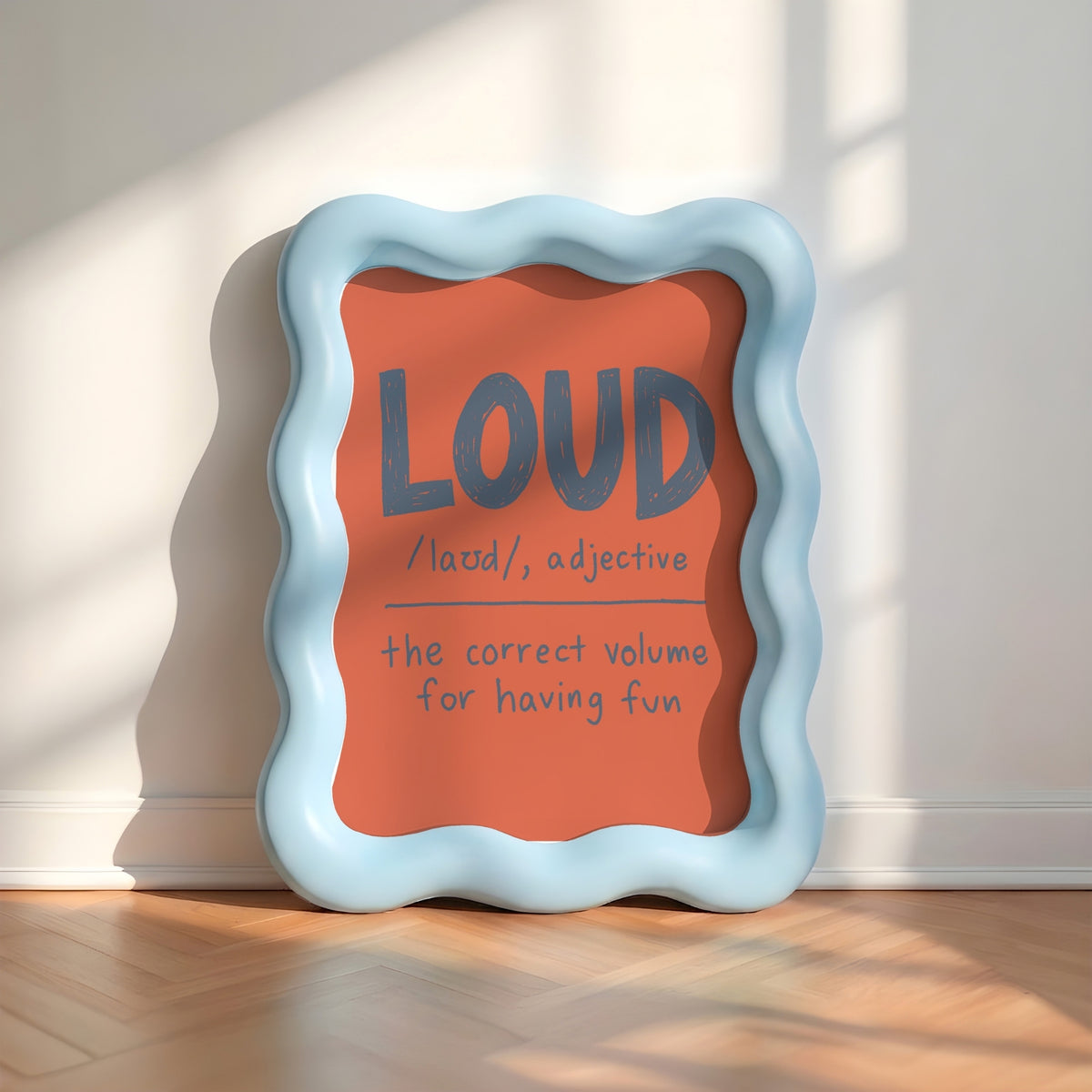 A decorative square sign with a wavy light blue frame leans against a white wall on a wood floor. The sign has a coral-orange center with the word “LOUD” in large gray letters and smaller handwritten-style text underneath reading “/lauid, adjective” and “the correct volume for having fun.