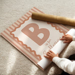 A person unrolls a decorative wall banner or poster on a beige carpet, with a brown cardboard mailing tube placed on top. The printed design features a large letter B, pink striped border, and the words 'is for Ballerina'.