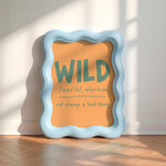 A wavy light blue picture frame leaning against a white wall on a wood floor, displaying an orange sign with the word 'WILD' and a dictionary-style definition.