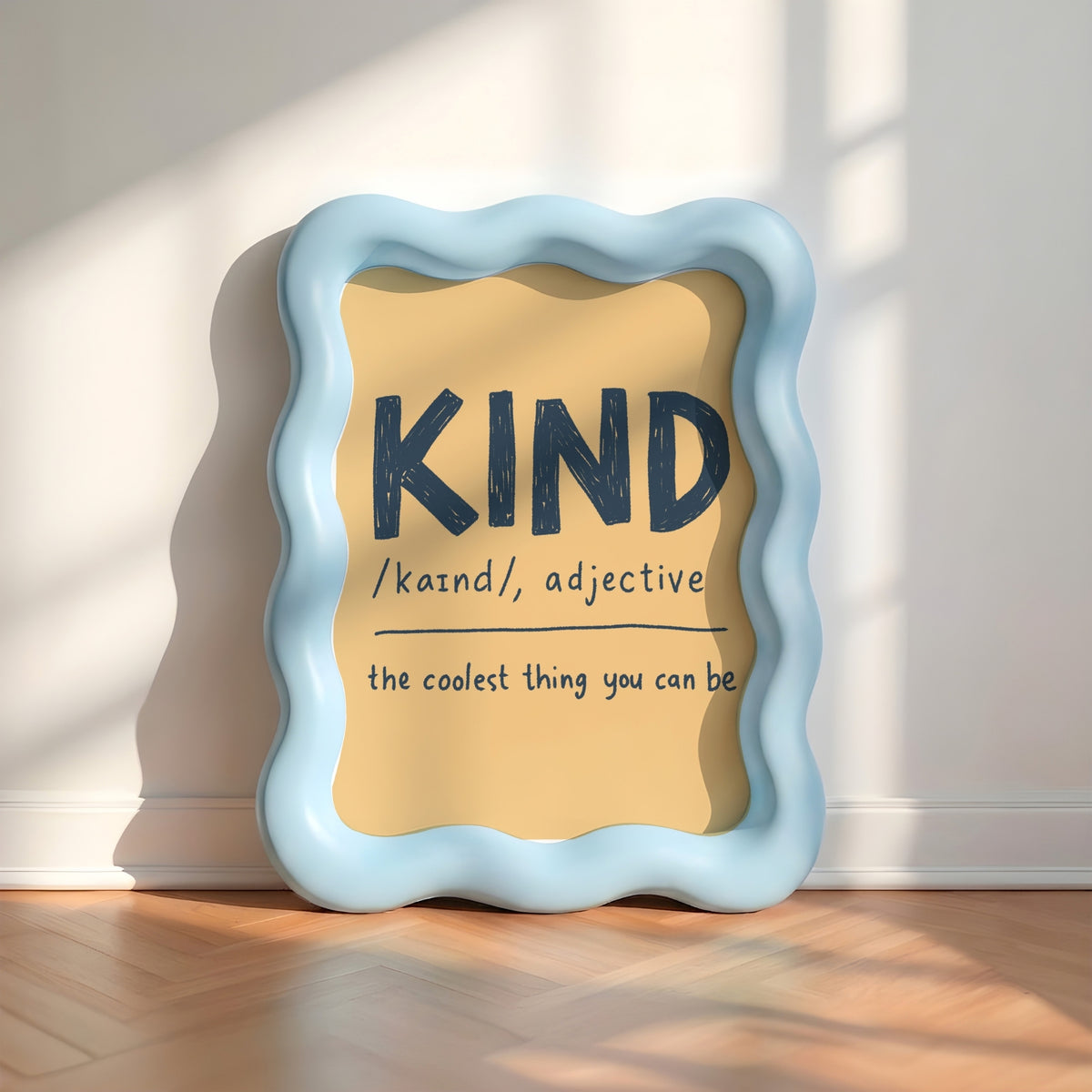 A decorative framed wall sign with a wavy light blue border and a warm beige insert, leaning against a white wall on a wood floor. The insert reads “KIND /kaind/, adjective” and “the coolest thing you can be.