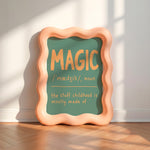 A decorative framed sign leaning against a wall on a wood floor, with a peach wavy frame and a green center that reads “MAGIC / ‘médÙÙk/, noun / the stuff childhood is mostly made of” in warm yellow text.