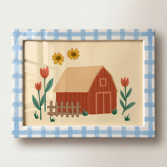 A framed decorative art print featuring a red barn, yellow flowers, and red tulips on a cream background, surrounded by a blue-and-white gingham border. The framed piece is photographed against a light neutral wall.