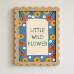 A framed wall art print with a scalloped light wood frame. The artwork features the text 'LITTLE WILD FLOWER' in green on a cream background, surrounded by a blue border decorated with yellow sunflowers, red poppies, and small white flowers.
