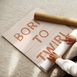 A person unrolls a sheet of wrapping paper on a beige carpet. The wrapping paper has thin pink stripes and large metallic rose-gold lettering that reads 'BORN TO TWIRL.