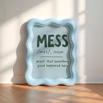 A light blue wavy-edged decorative sign leaning against a white wall on a wooden floor, with printed text reading “MESS /mes/, noun proof that something good happened here” in dark green and black lettering.