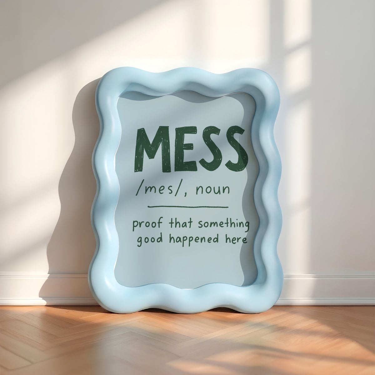 A light blue wavy-edged decorative sign leaning against a white wall on a wooden floor, with printed text reading “MESS /mes/, noun proof that something good happened here” in dark green and black lettering.