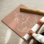 A person's hands are shown unrolling a large sheet of dusty rose pink vinyl floor decals with a white line-art design of a ballerina, next to a roll of matching pink decals on a beige carpet.