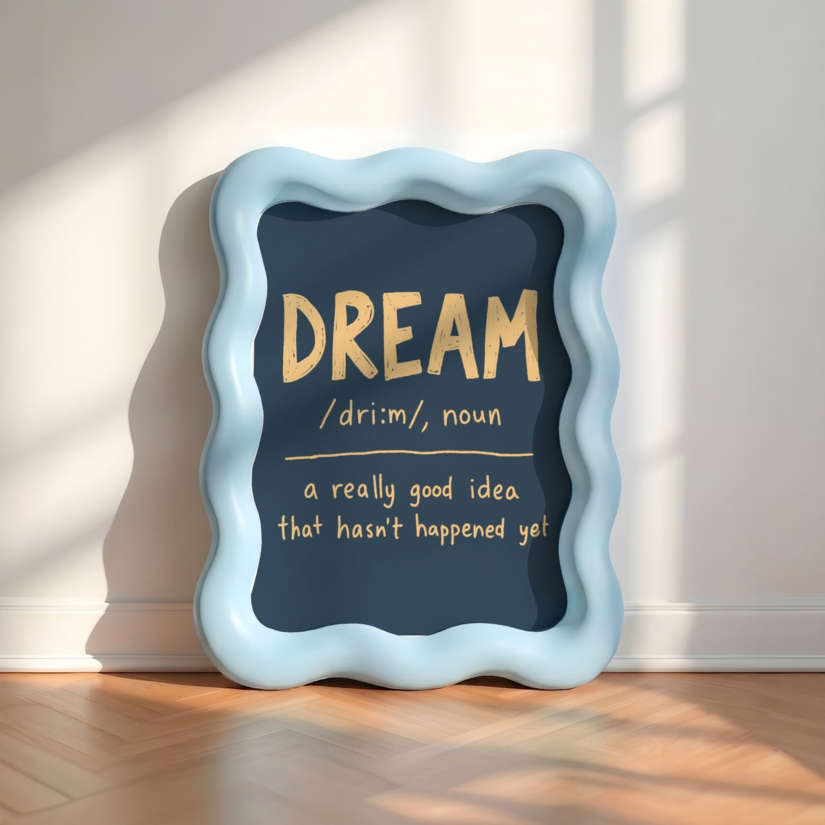 A decorative framed wall sign with a wavy light blue border and a dark navy center panel, leaning against a white wall on a wood floor. The panel displays the word “DREAM” in large gold letters with a dictionary-style definition below in white and gold text.