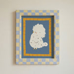 A framed art print featuring a cute white fluffy bird with small gray spots on a dusty blue background, surrounded by a yellow-orange patterned border and a blue-and-beige checkered frame, mounted on a light beige wall.